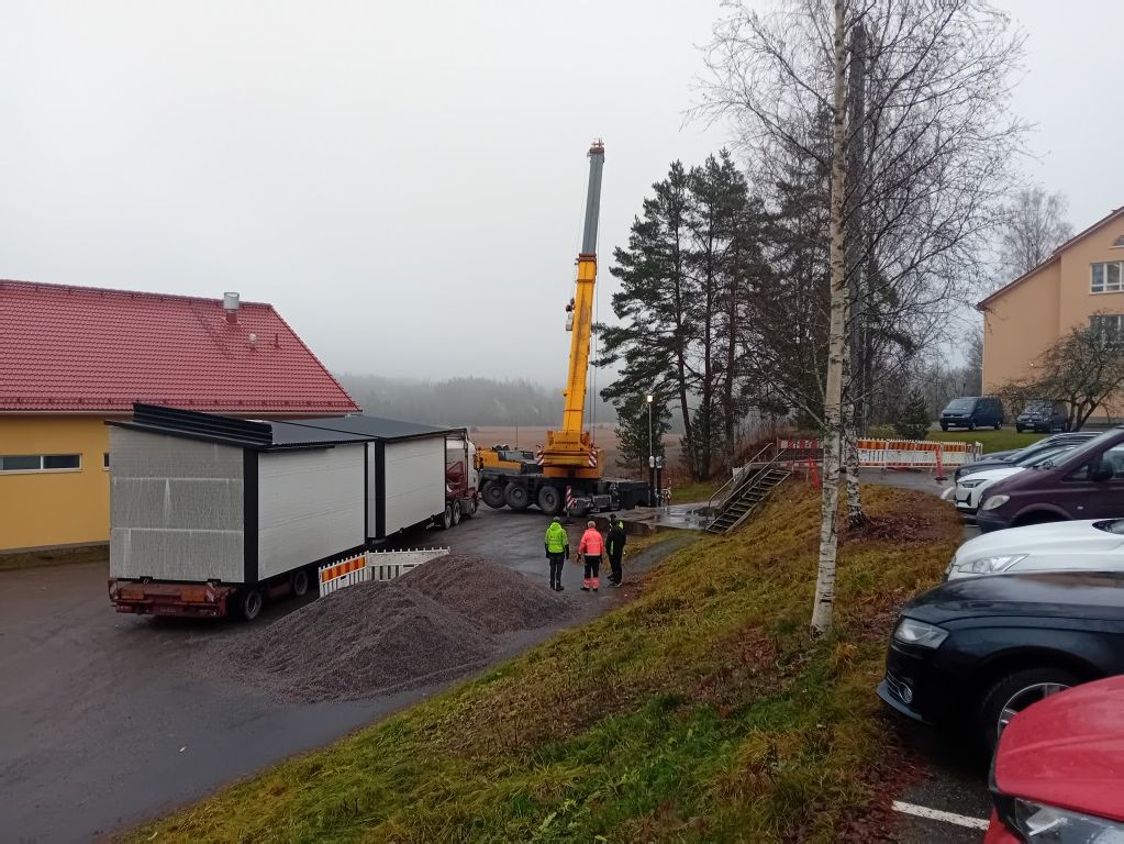 Sumuinen päivä, jossa näkyy keltainen nosturi, kaksi siirrettävää rakennusta, kasa soraa ja kolme työntekijää. Taustalla on rakennuksia ja autoja.