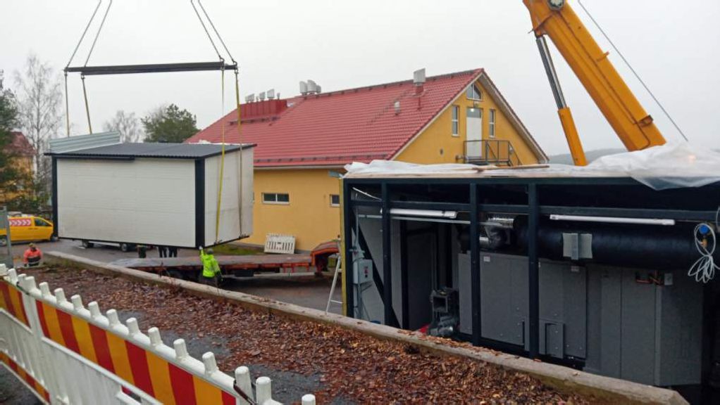 Ulkona nostetaan valkoista siirrettävää rakennusta nosturilla, taustalla keltainen rakennus punaisella katolla ja työmaa-aita.